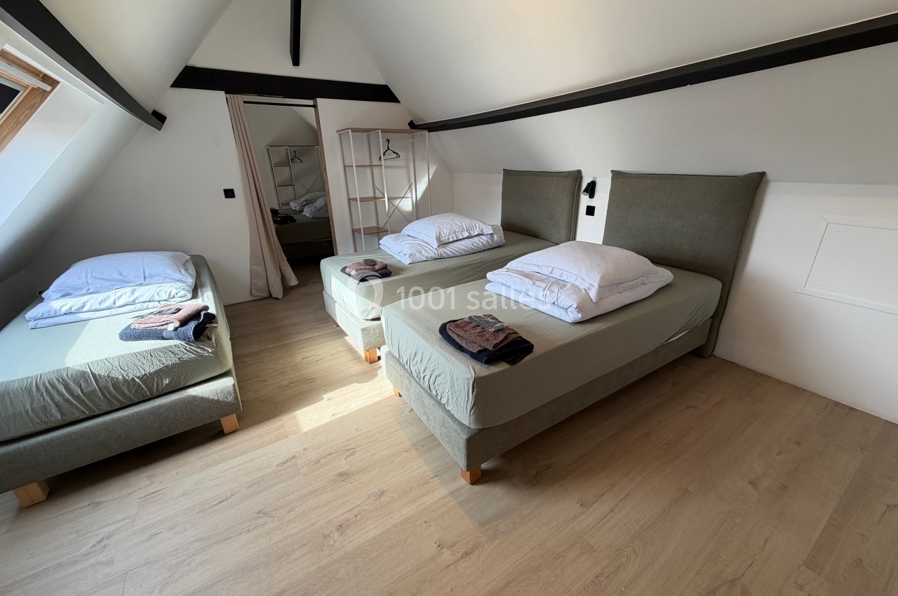 Chambre mansardée avec trois lits simples, parquet clair, poutres apparentes et lumière naturelle provenant d'une fenêtre.