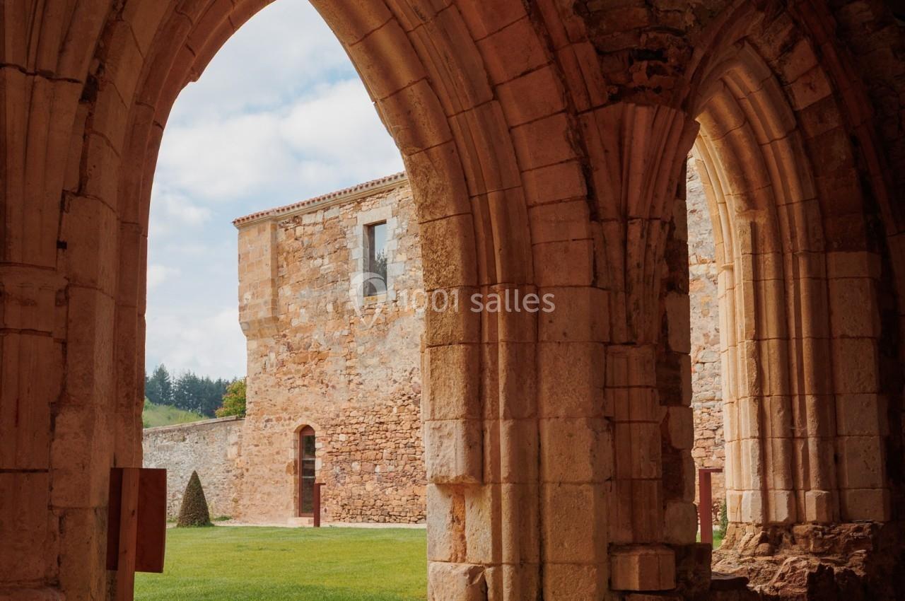 Vue depuis des arcs en pierre d'une cour herbeuse entourée de bâtiments anciens en pierre sous un ciel partiellement nuageux.