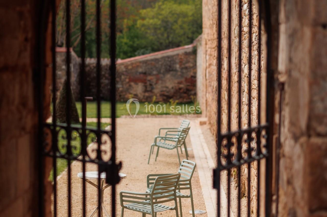 Vue à travers une porte en fer forgé ouverte sur une terrasse avec des chaises en métal et un jardin en arrière-plan.