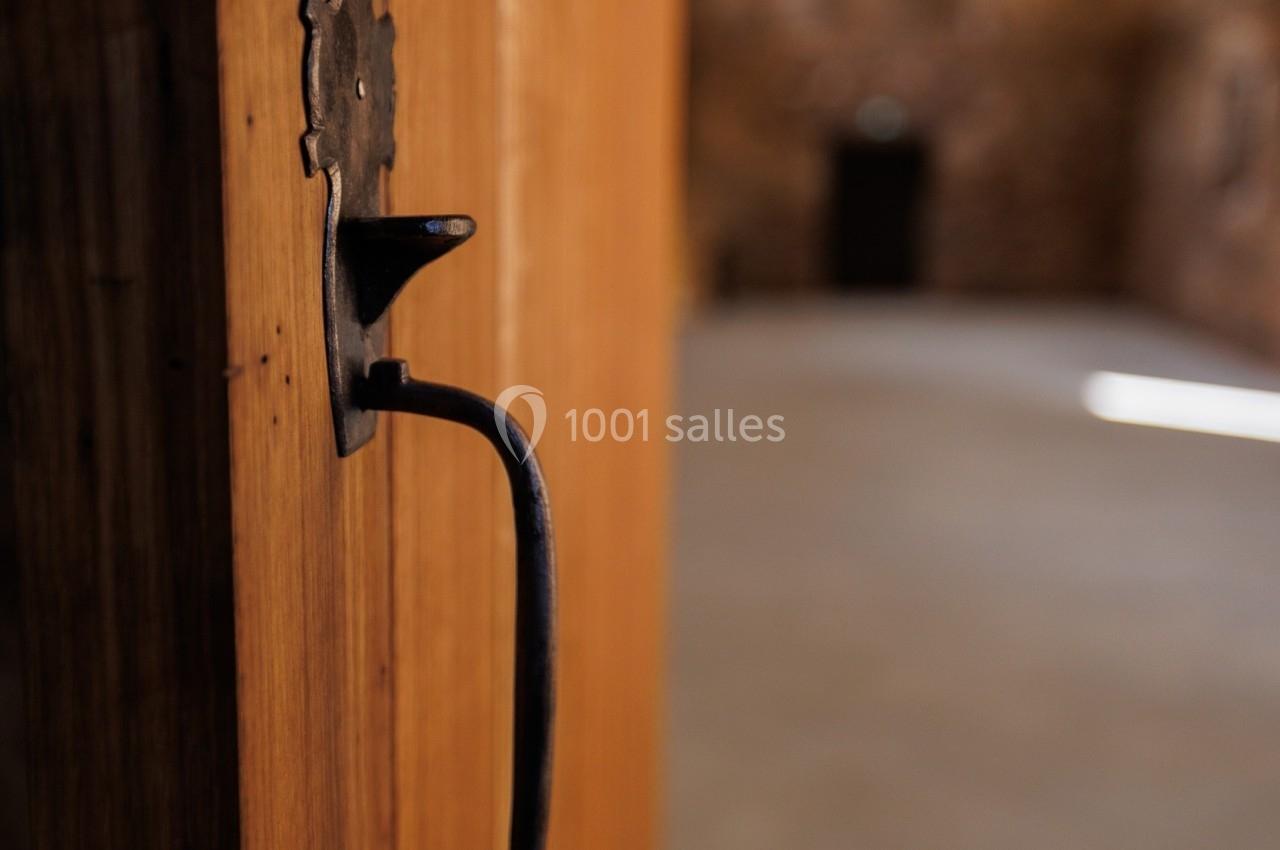 Porte en bois entrouverte avec une poignée en métal noir, donnant sur une pièce lumineuse aux murs en pierre.