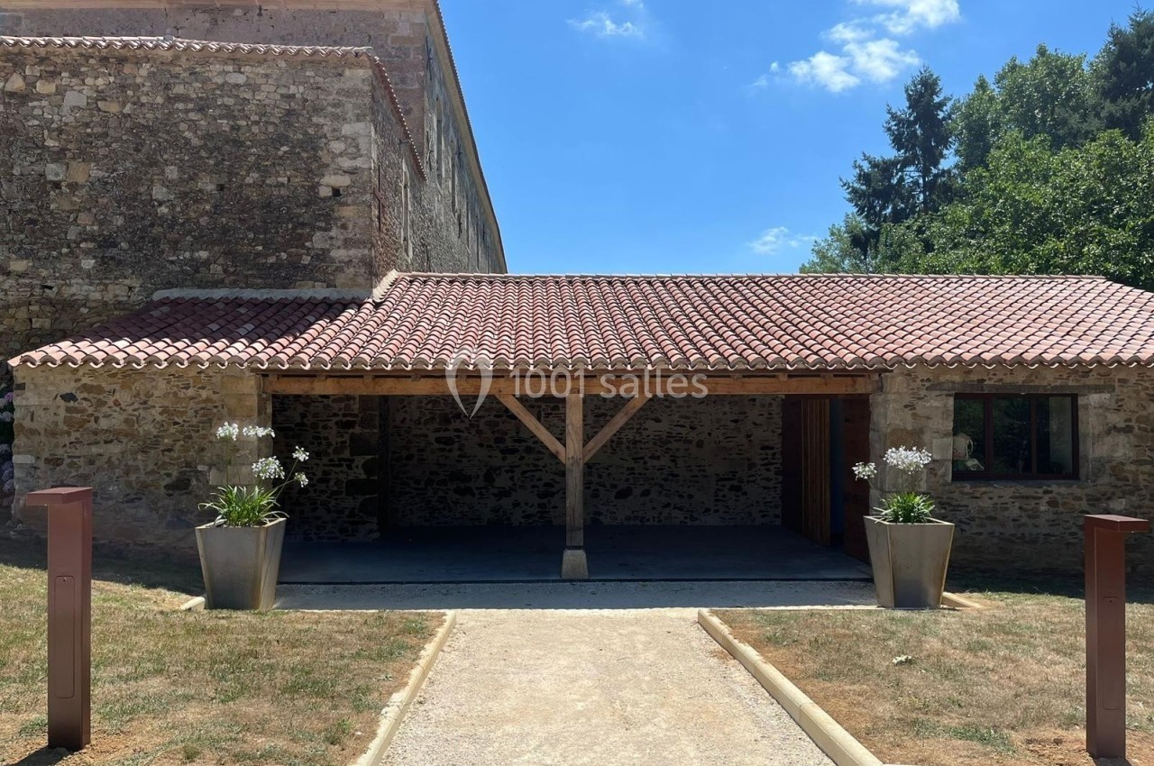 Bâtiment en pierre avec toit en tuiles, abri ouvert en bois et deux grandes jardinières contenant des fleurs blanches.