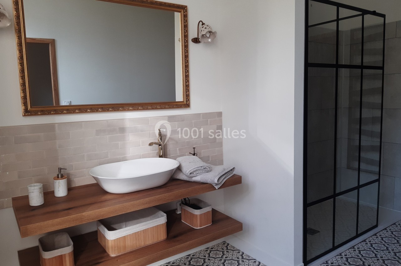 Salle de bain moderne avec vasque sur meuble en bois, miroir encadré doré et douche à paroi vitrée noire.