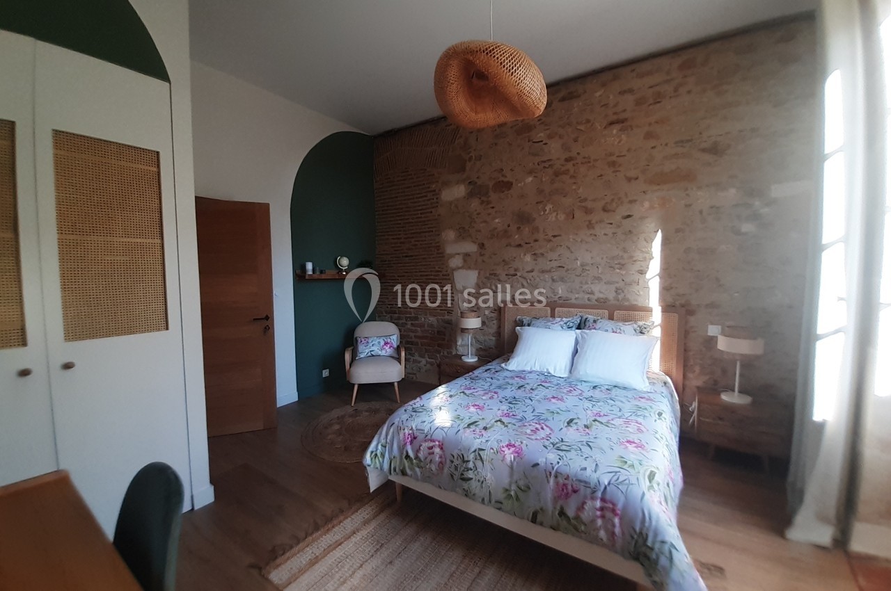 Chambre lumineuse avec un mur en pierre, lit double, mobilier en bois et fauteuil près d'une étagère.