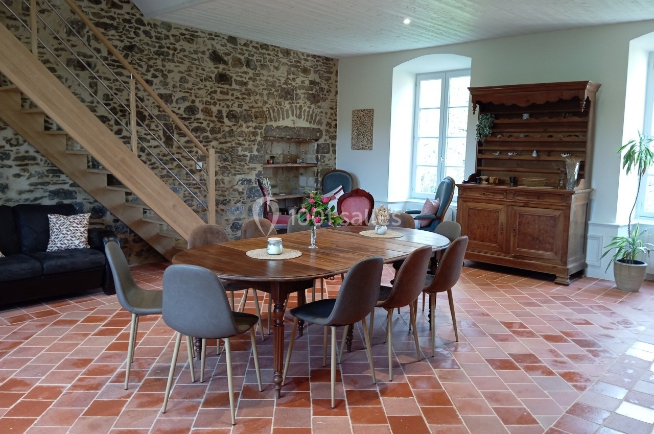 Salle à manger lumineuse avec table en bois, chaises modernes, escalier en bois, murs en pierre et carrelage au sol.