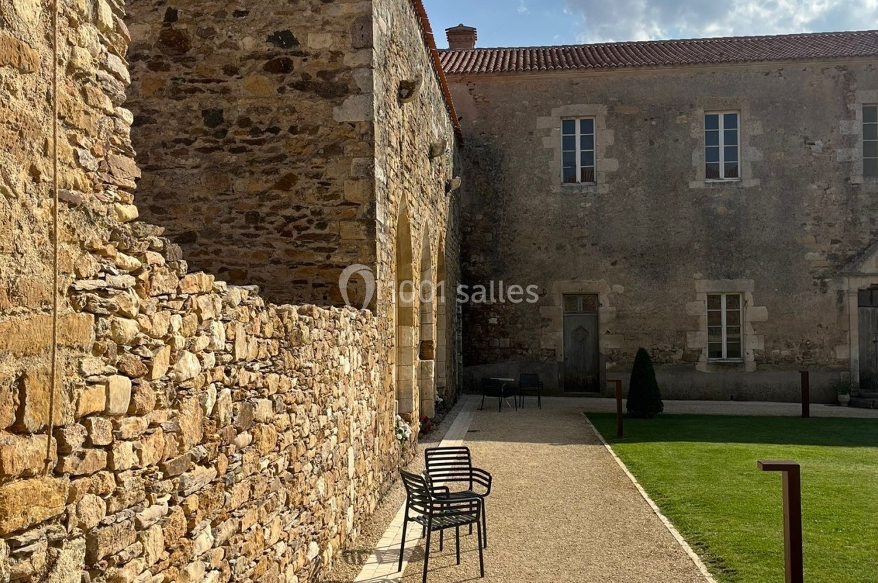 Allée pavée bordée de bancs en métal, longeant un bâtiment en pierre sous un ciel partiellement nuageux.