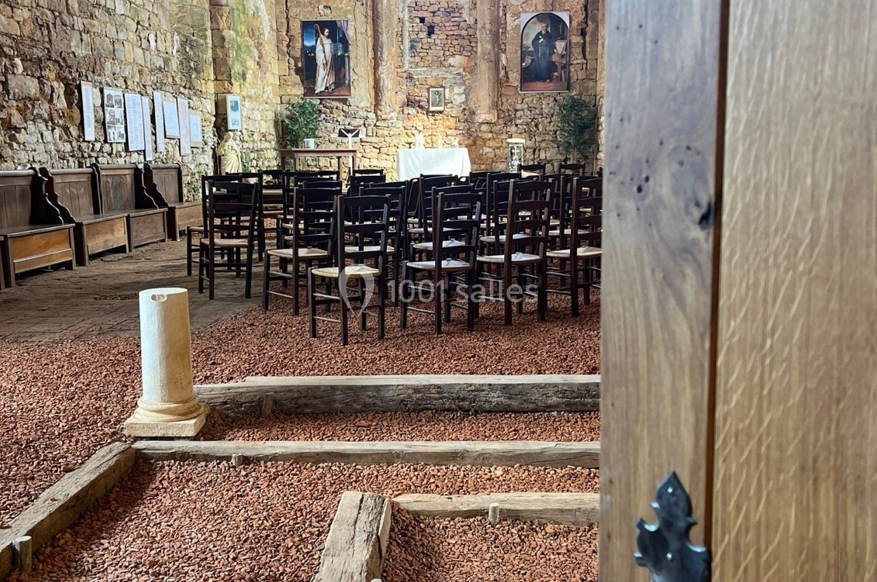 Intérieur d'une chapelle en pierre avec des rangées de chaises, un autel au fond et une porte en bois entrouverte au premier…