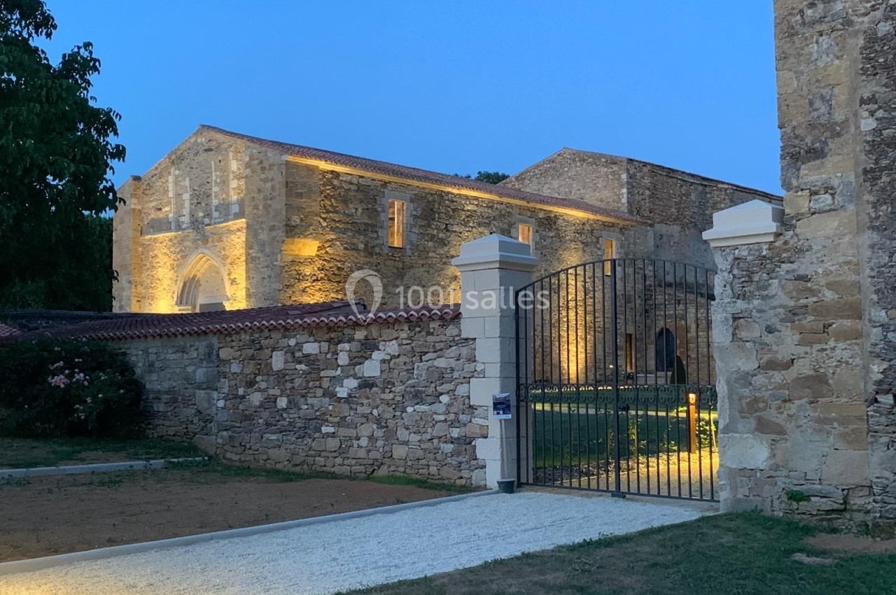 Vue d'un bâtiment en pierre éclairé de nuit, entouré d'un mur avec une grille en fer forgé ouverte.