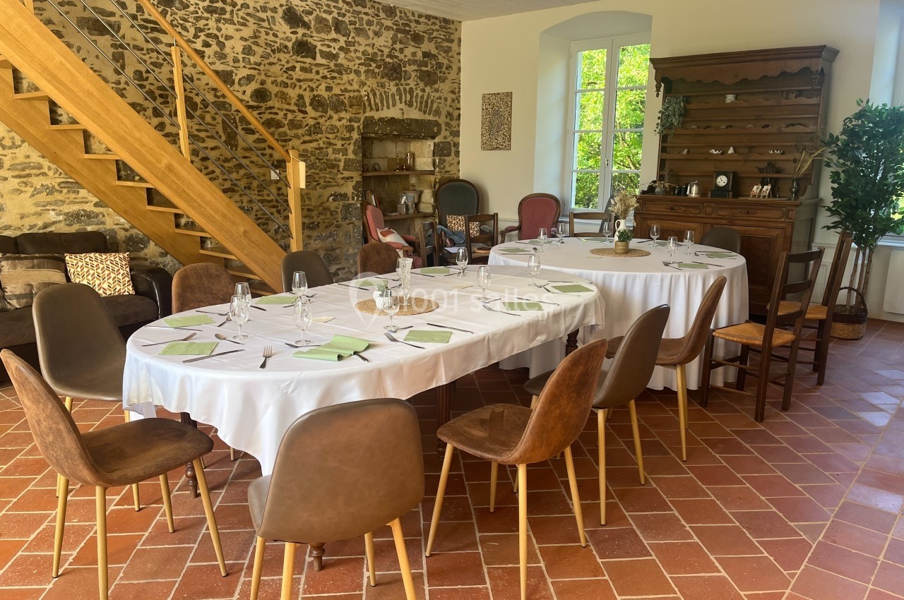 Salle à manger lumineuse avec tables dressées, chaises variées, escalier en bois et mur en pierre apparente.