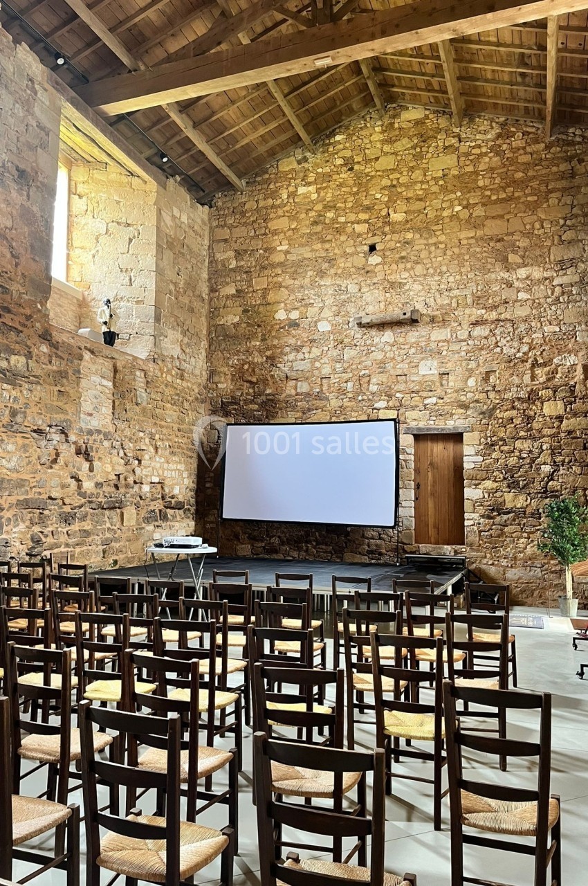 Salle en pierre avec un écran de projection, des rangées de chaises en bois et un plafond en poutres apparentes.