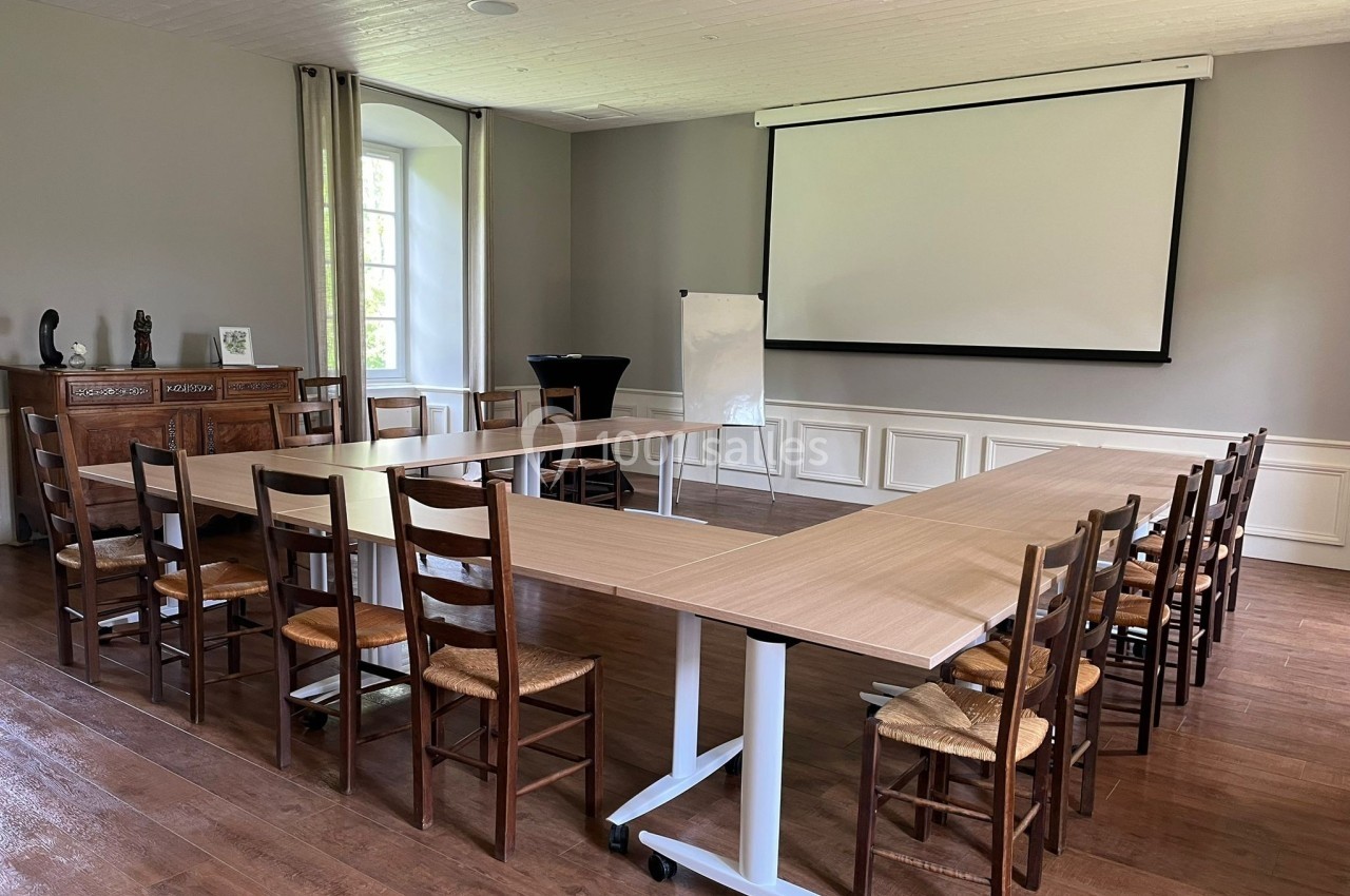Salle de réunion lumineuse avec tables en U, chaises en bois, tableau blanc et écran de projection.