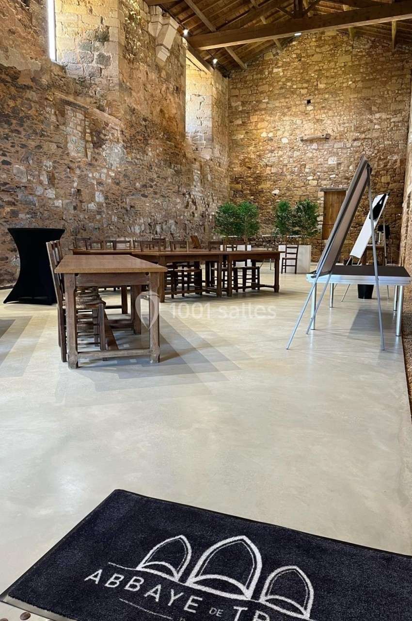 Salle en pierre avec tables en bois, tableau de présentation et tapis marqué ’Abbaye de Trizay’.