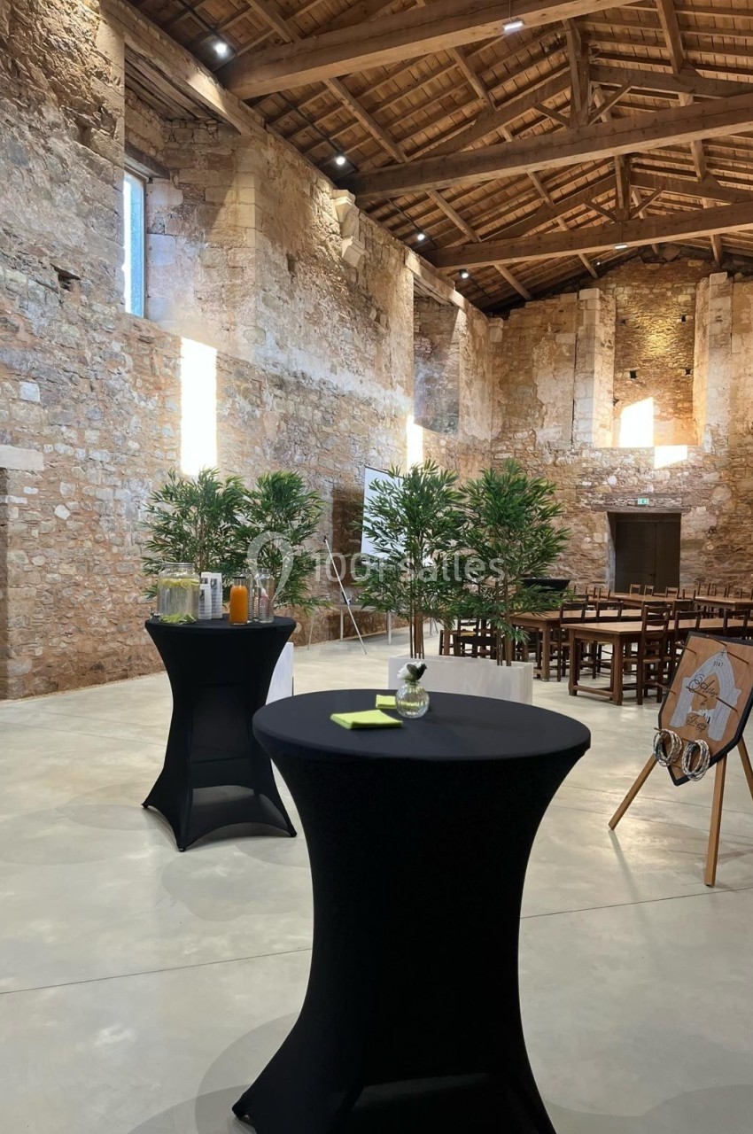 Salle en pierre avec tables hautes noires, boissons, plantes décoratives et chaises disposées pour un événement.