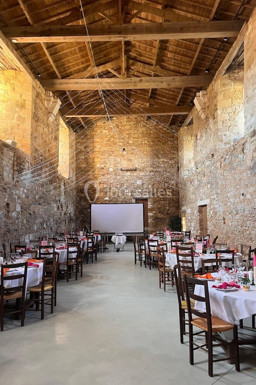 Salle en pierre avec tables dressées pour un événement, éclairée par des guirlandes lumineuses suspendues.