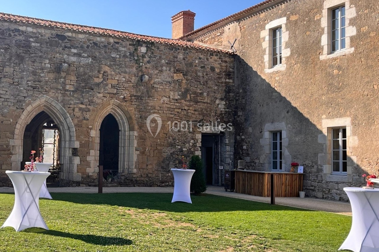 Cour extérieure d'un bâtiment en pierre ancienne avec des tables hautes blanches et un bar en bois.