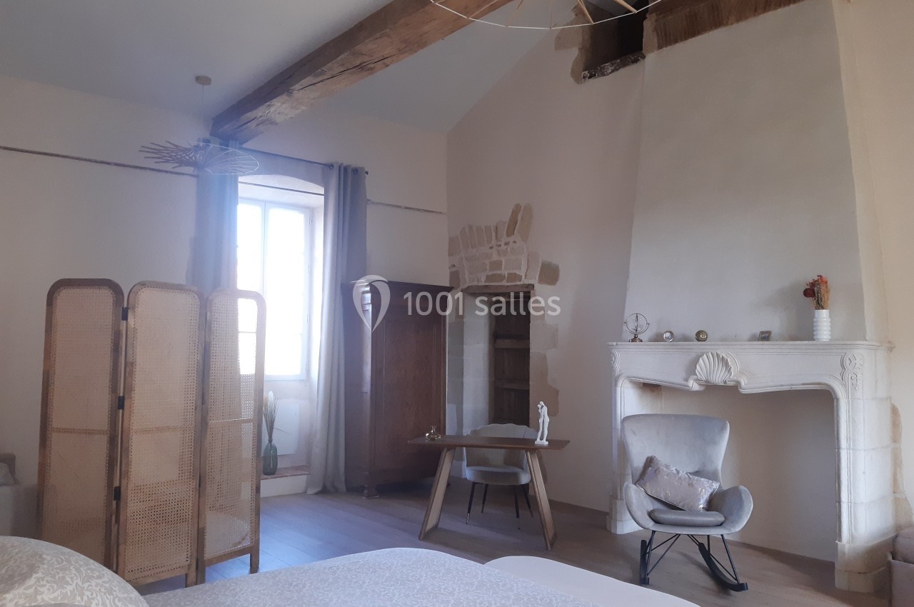 Chambre lumineuse avec poutres apparentes, cheminée en pierre, paravent en rotin et mobilier moderne.