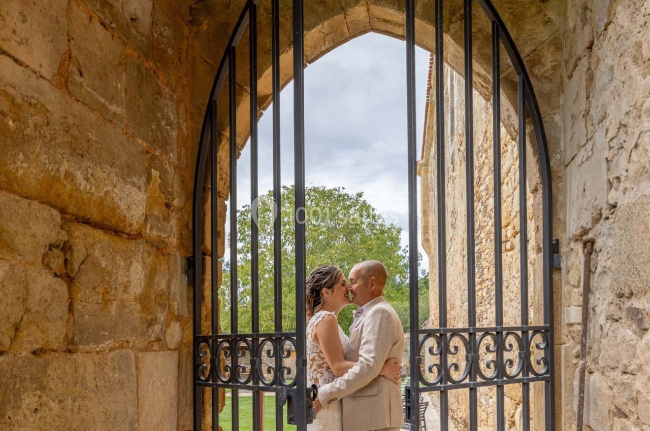 Un couple s'embrasse devant une porte en fer forgé dans un cadre en pierre, avec un arbre visible en arrière-plan.