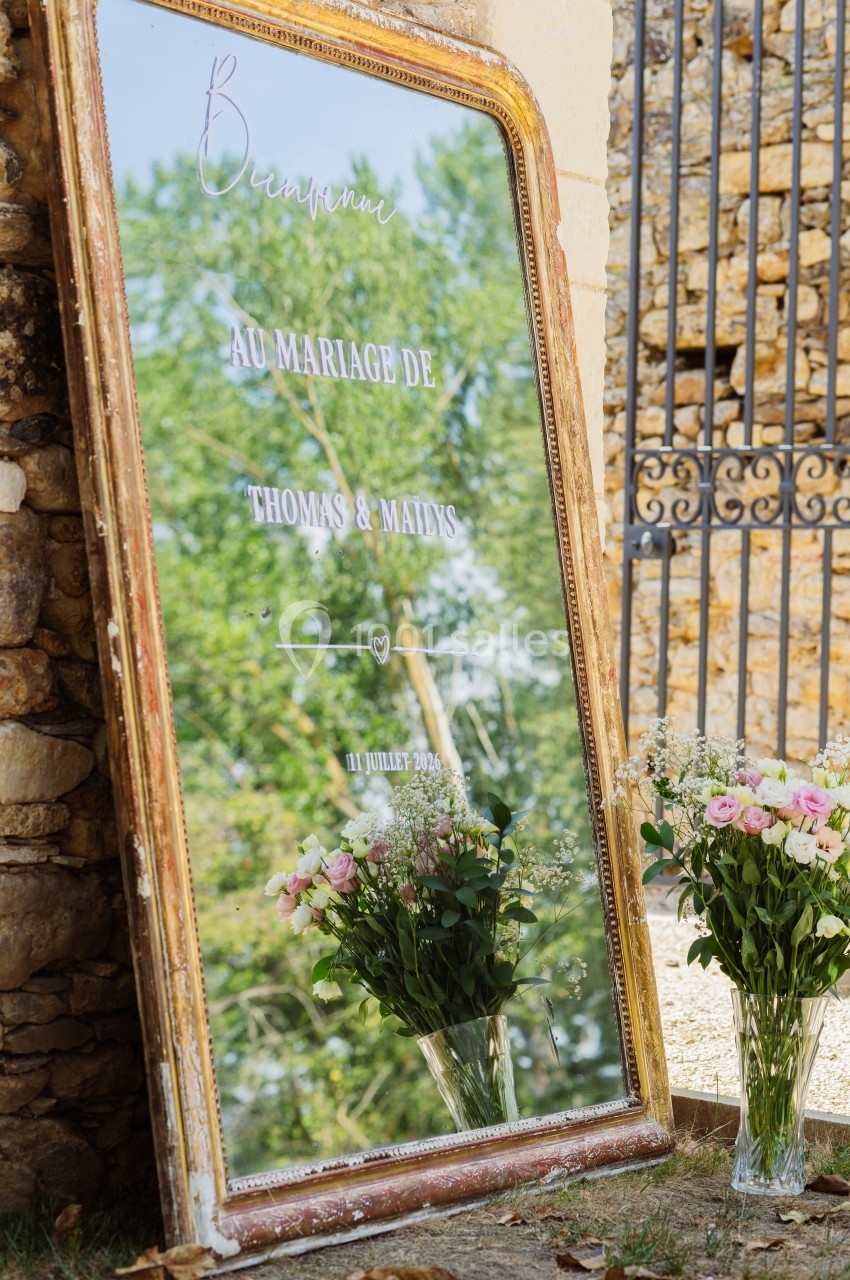 Grand miroir doré avec un message de bienvenue pour un mariage, entouré de fleurs roses et blanches.