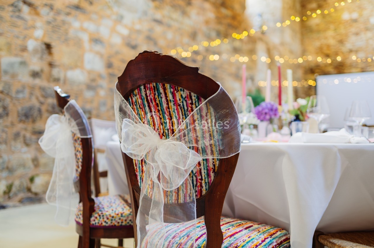 Chaise en bois décorée d'un nœud en tissu blanc, placée près d'une table dressée dans une salle en pierre éclairée.