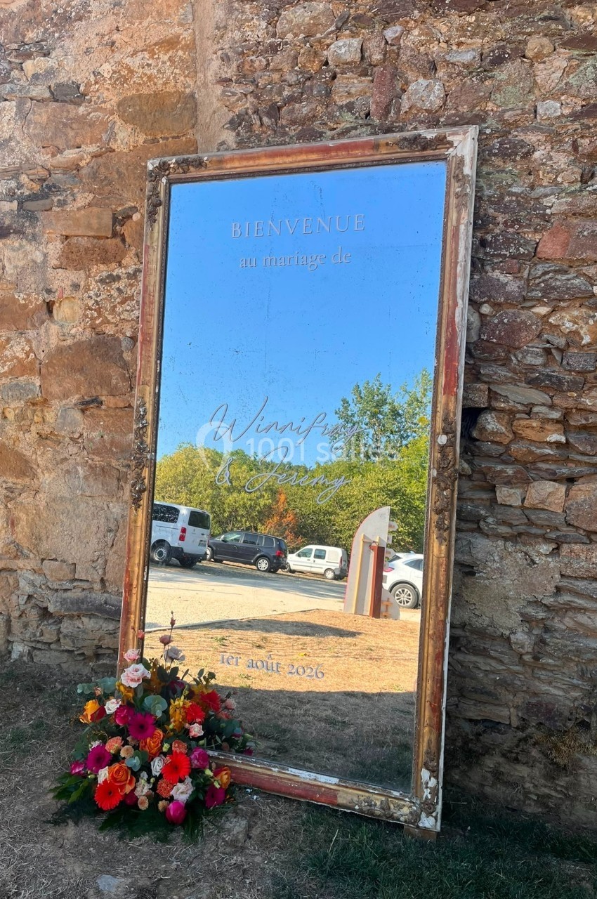 Miroir ancien appuyé contre un mur en pierre, affichant un message de bienvenue pour un mariage, avec un bouquet de fleurs…
