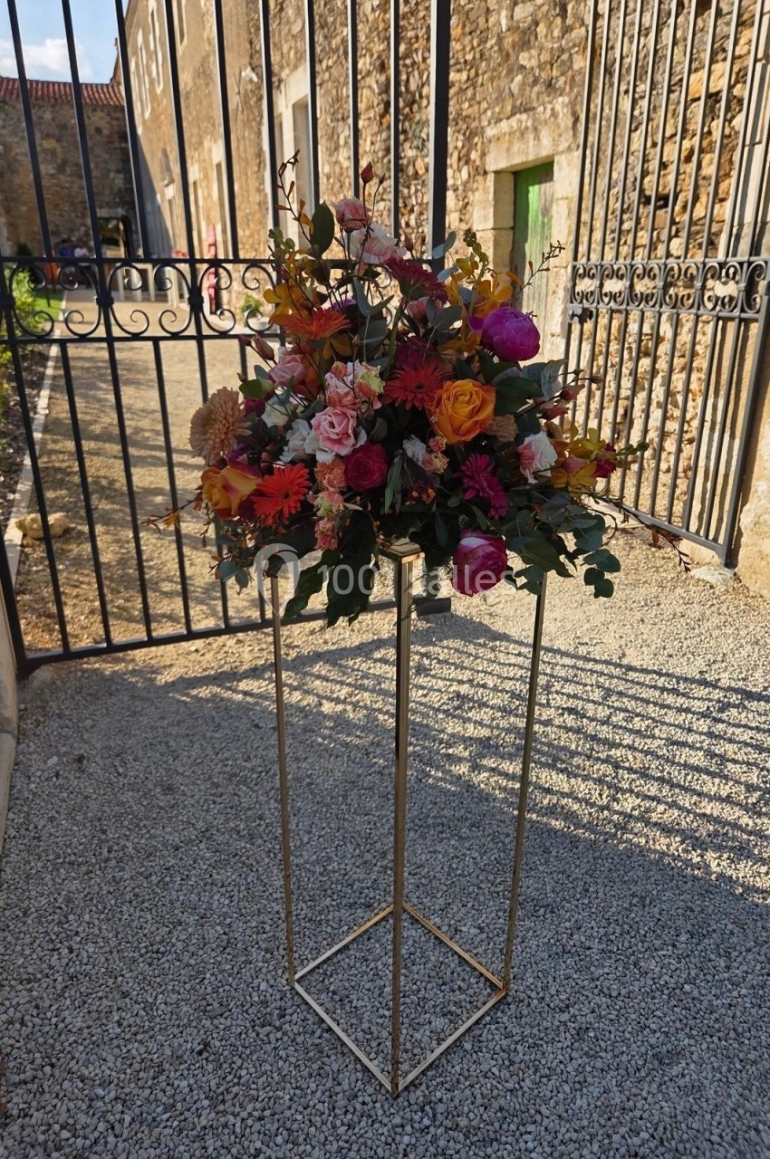 Arrangement floral coloré sur un support métallique doré, placé devant une grille en fer forgé et un mur en pierre.