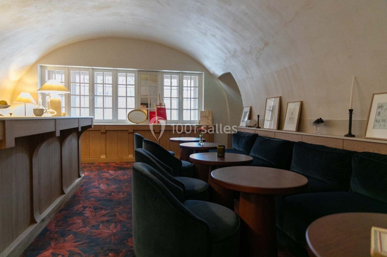 Salle voûtée avec canapés en velours, tables en bois, éclairage doux et fenêtres laissant entrer la lumière naturelle.