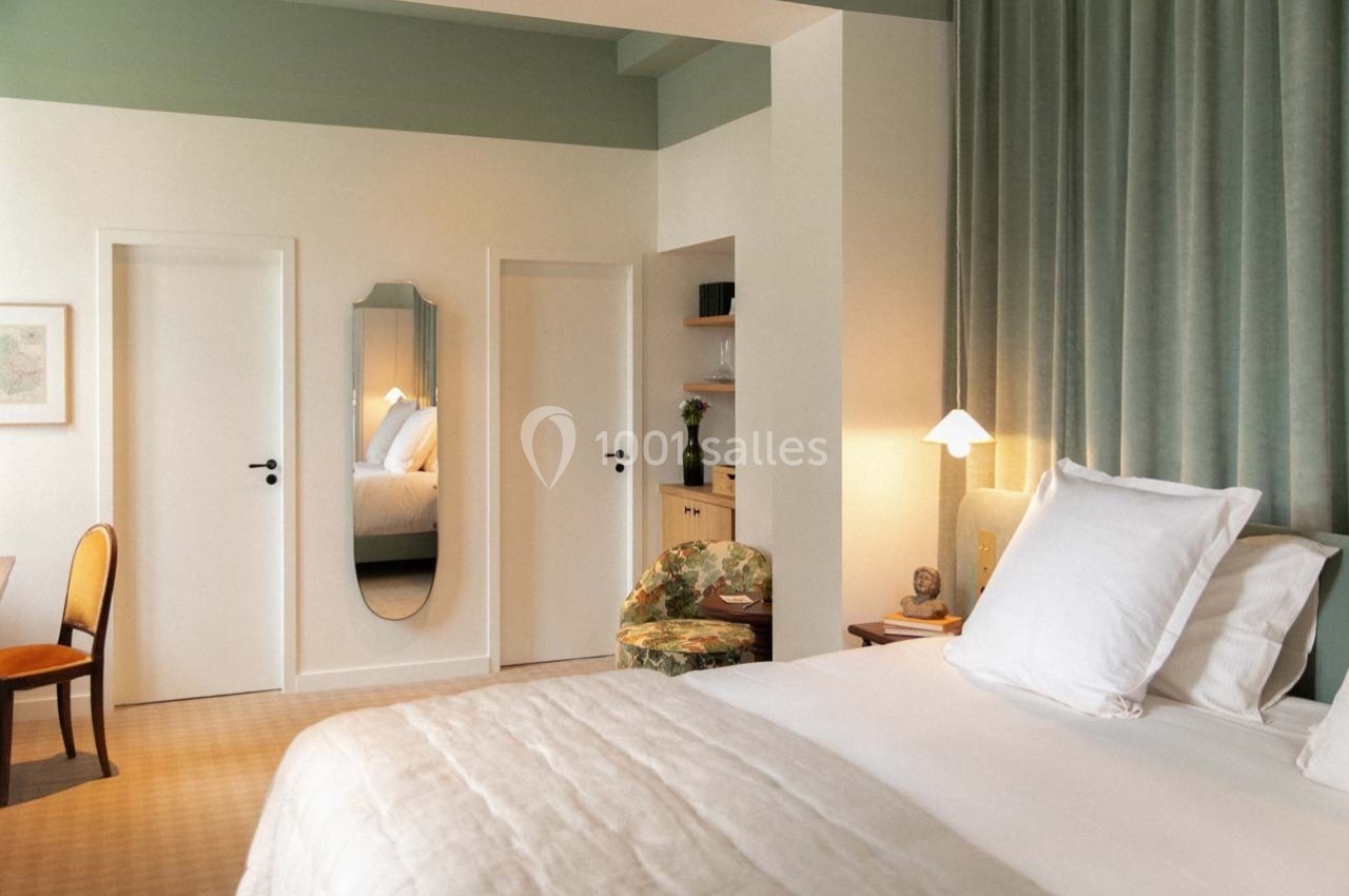 Chambre lumineuse avec lit double, miroir mural, rideaux verts, bureau en bois et fauteuil fleuri.