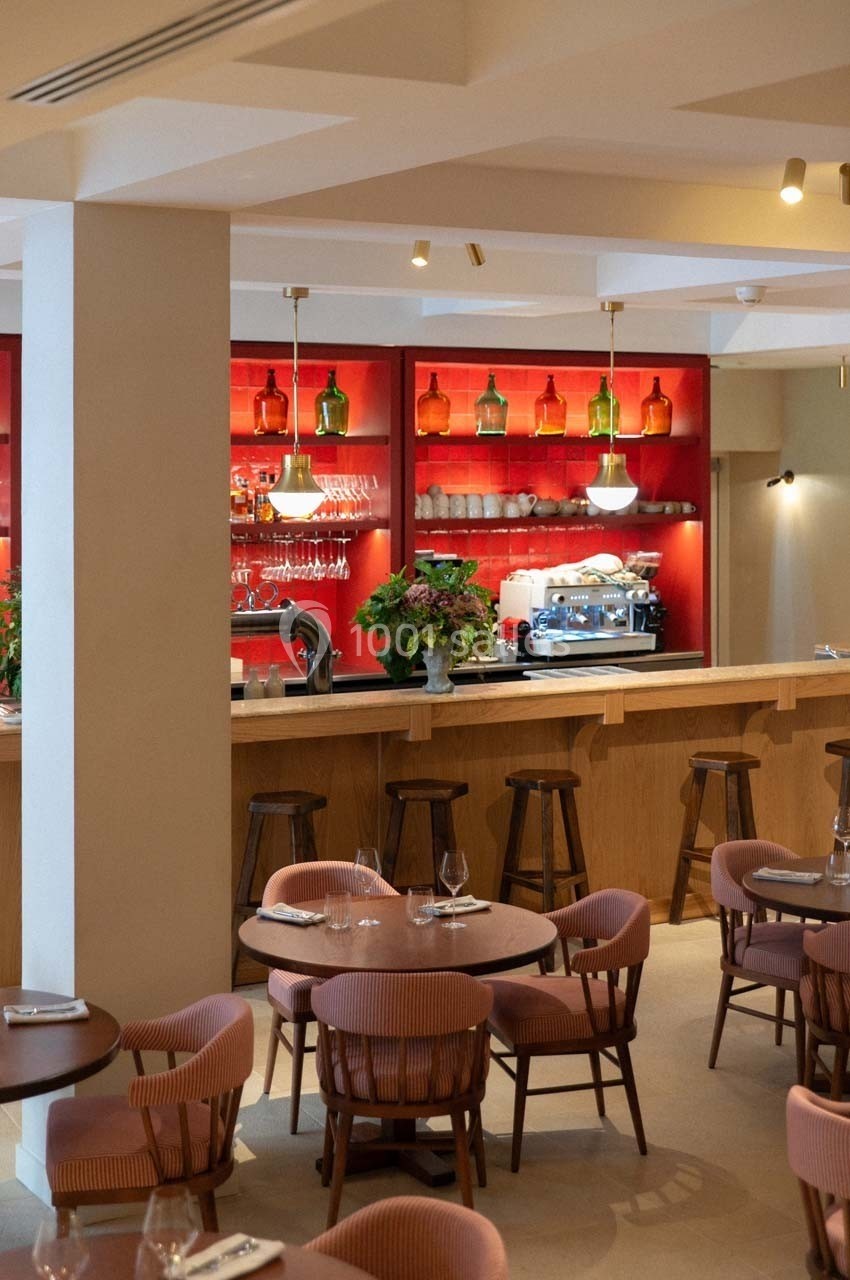 Salle de restaurant avec tables dressées, chaises en bois et un bar en arrière-plan avec étagères rouges et éclairage…