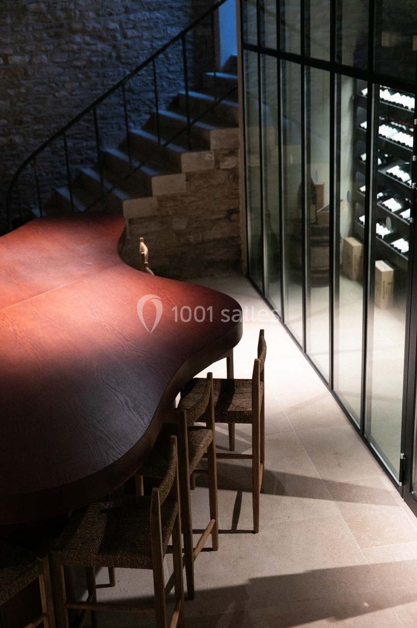 Salle moderne avec une grande table en bois, des chaises et une cave à vin visible derrière une paroi vitrée.