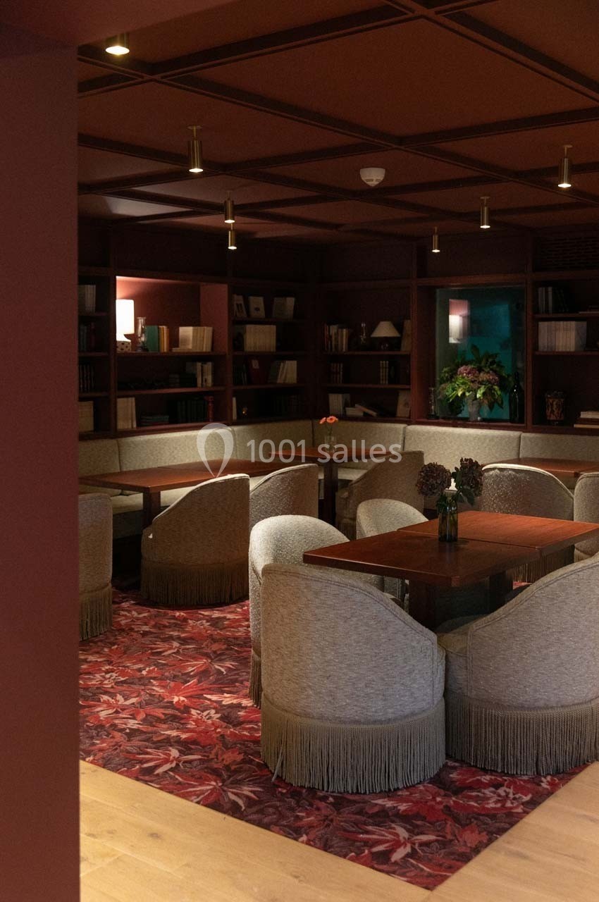 Salle de lecture chaleureuse avec fauteuils en tissu, tables en bois, étagères remplies de livres et éclairage tamisé.
