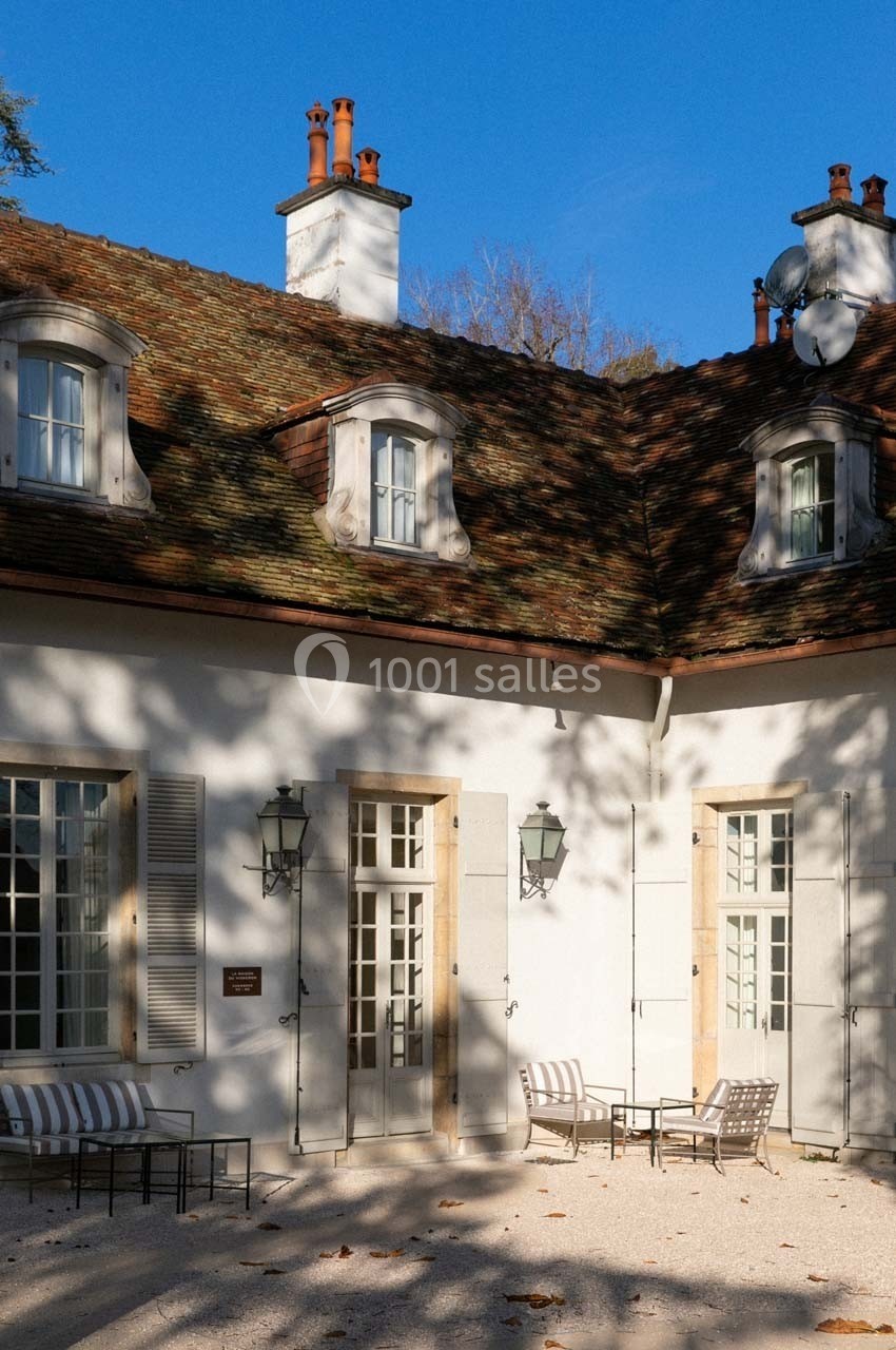 Façade d'une maison avec toit en tuiles, fenêtres mansardées et terrasse équipée de chaises sous un ciel bleu.