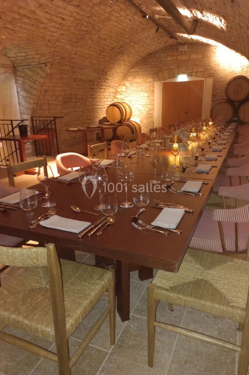 Grande table en bois dressée avec vaisselle et verres, située dans une cave voûtée avec des tonneaux en arrière-plan.