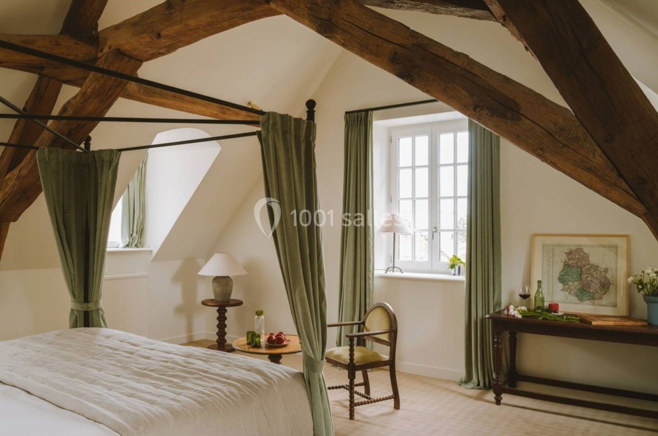 Chambre lumineuse avec poutres apparentes, lit à baldaquin, table en bois, chaise et fenêtre encadrée de rideaux verts.