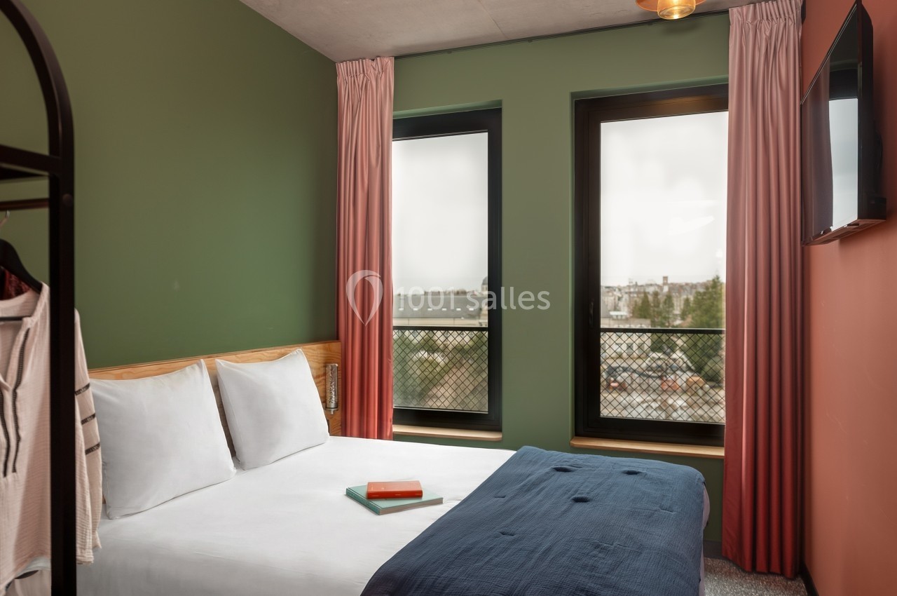 Chambre lumineuse avec lit double, murs verts et rouges, grandes fenêtres donnant sur un paysage urbain.