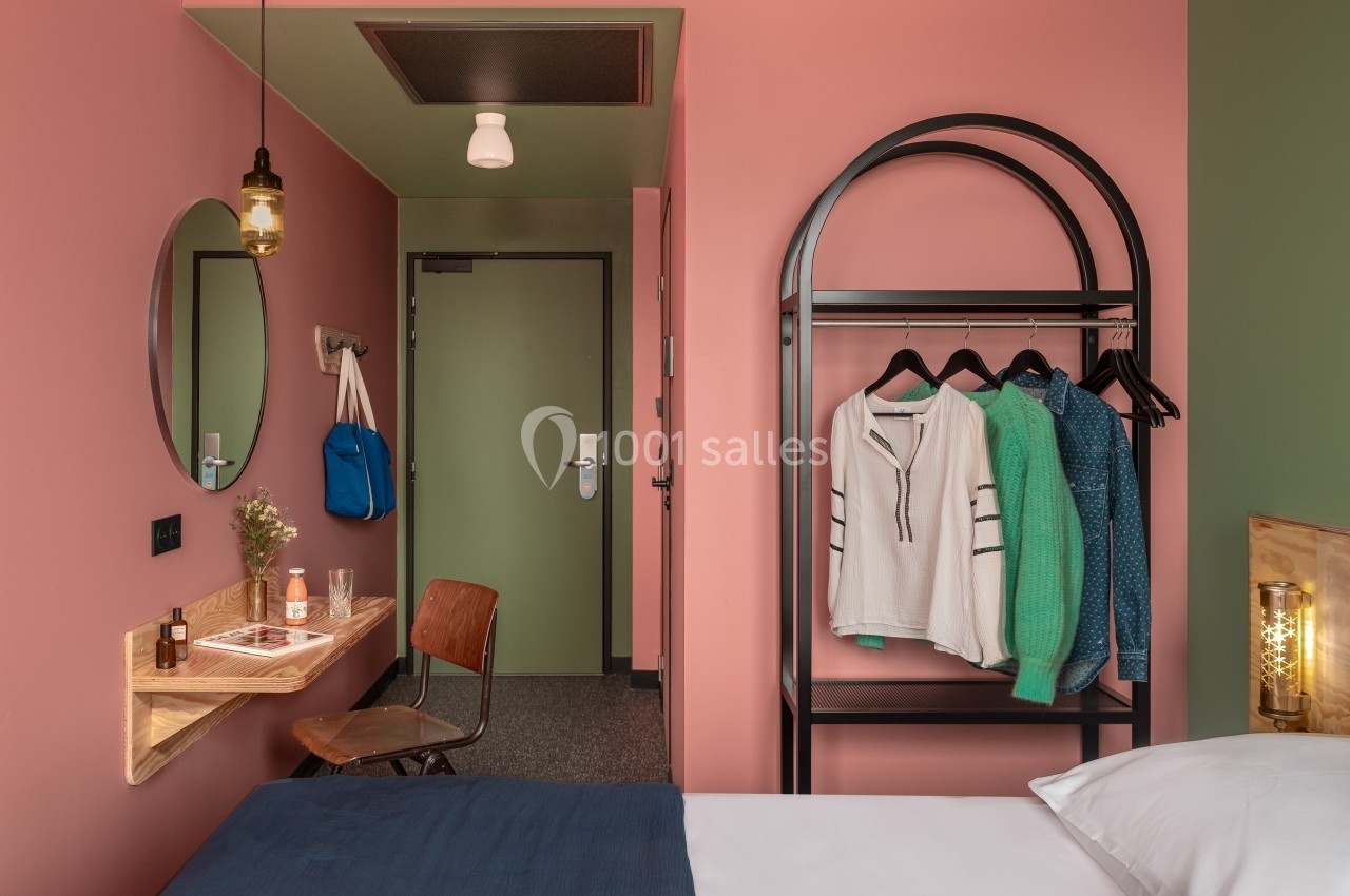 Chambre d'hôtel avec murs roses, lit, bureau minimaliste, porte verte et portant avec vêtements suspendus.