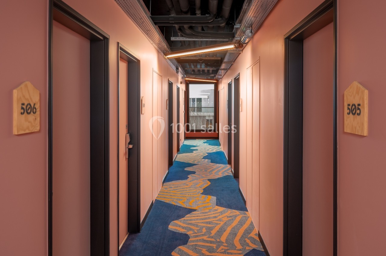 Couloir d'hôtel avec murs roses, moquette bleue à motifs et portes numérotées de chaque côté.