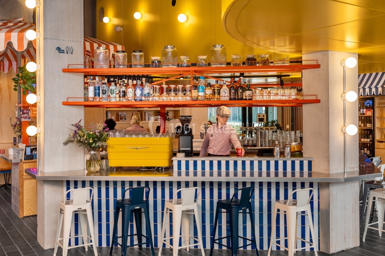 Bar coloré avec comptoir rayé bleu et blanc, étagères orange, décorations lumineuses et une personne servant une boisson.