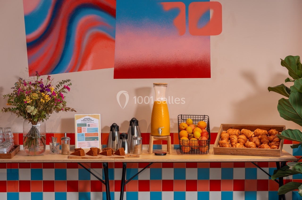 Table avec petit-déjeuner comprenant croissants, oranges, jus, café, fleurs et décorations colorées en arrière-plan.