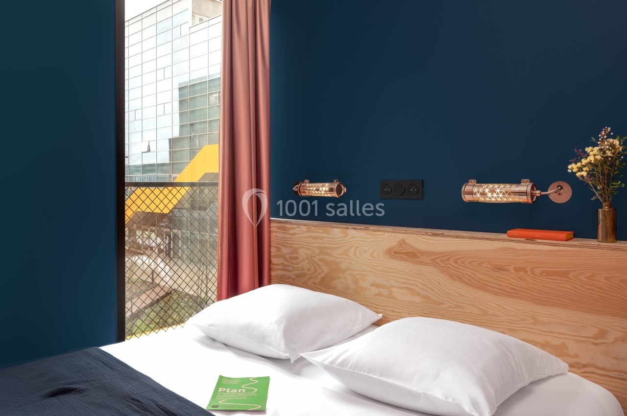 Lit avec tête de lit en bois clair, mur bleu, lampes murales cuivrées, fenêtre avec vue sur un bâtiment moderne.