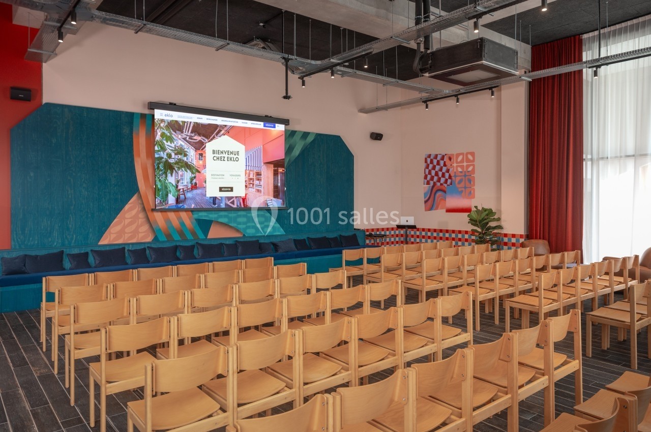 Salle de conférence moderne avec des rangées de chaises en bois, un écran de projection et une décoration colorée.