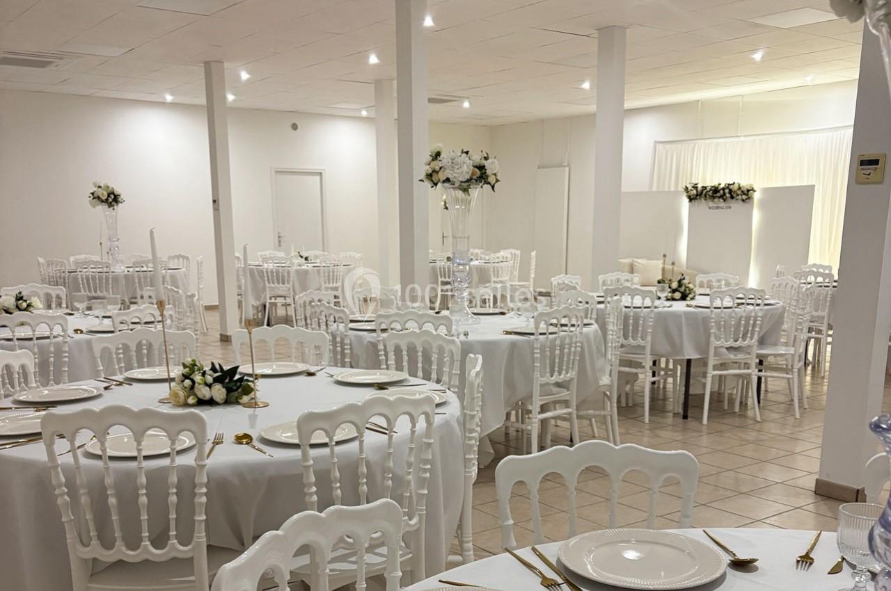 Salle de réception lumineuse avec tables rondes dressées, chaises blanches et décorations florales élégantes.