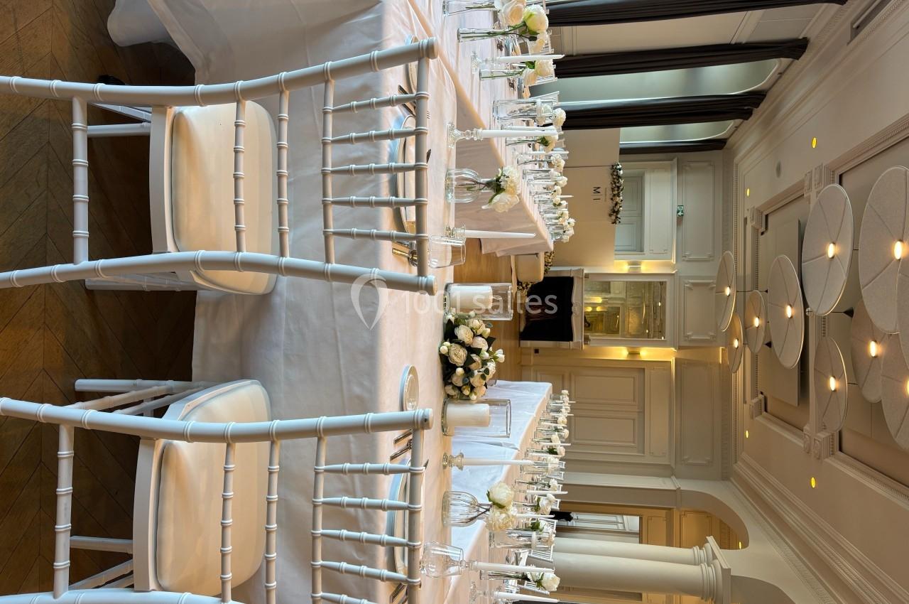 Salle élégante avec tables dressées, nappes blanches, chaises blanches et décor floral dans un cadre lumineux.