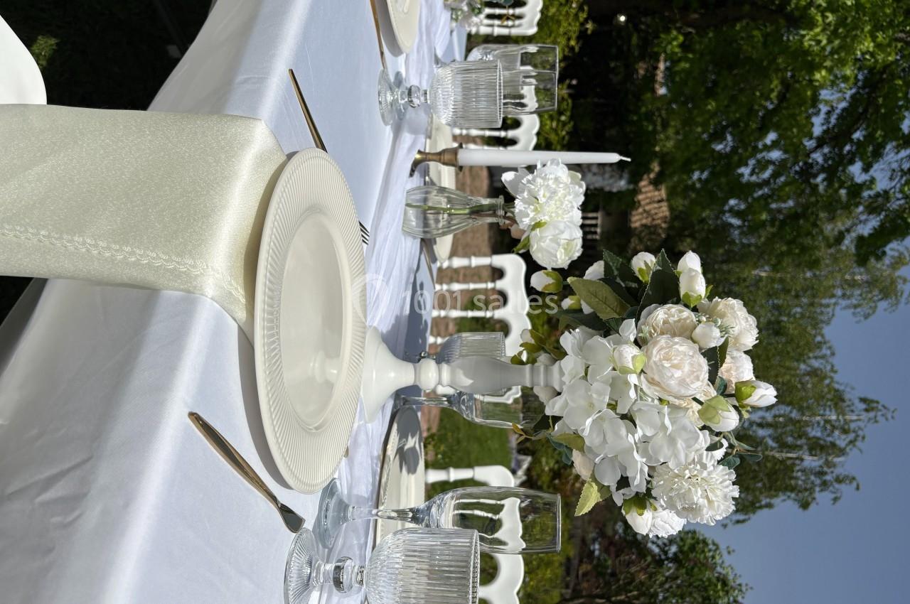 Table dressée en extérieur avec nappes blanches, vaisselle élégante et centre de table floral sous un ciel dégagé.