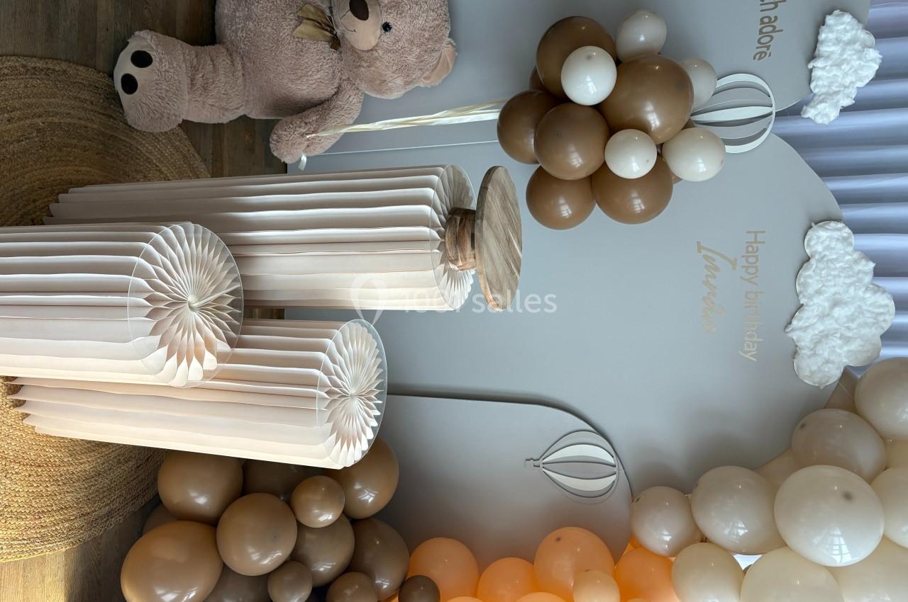 Décoration d'anniversaire avec ballons beige et blanc, ours en peluche et colonnes en papier plissé sur fond neutre.