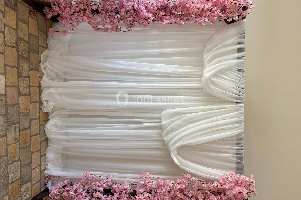 Rideau blanc drapé encadré de branches de fleurs roses, installé devant un mur clair et un sol en pierre.