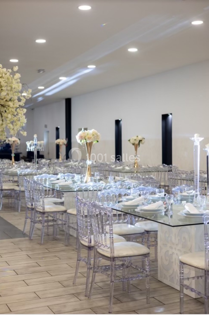 Salle de réception élégante avec tables dressées, chaises transparentes et décorations florales blanches.