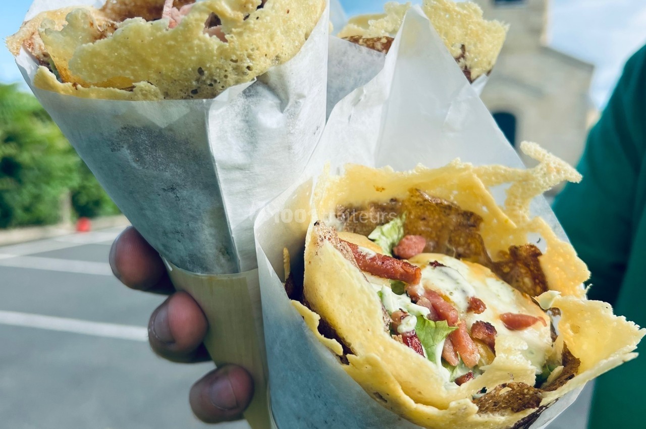 Deux cônes de galettes croustillantes garnies de salade, fromage et lardons, tenus à la main en extérieur.