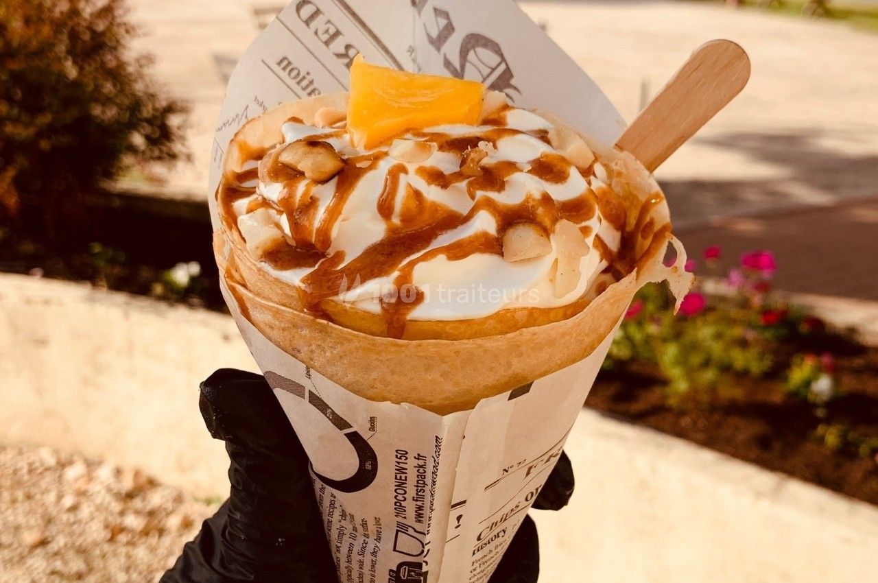 Crêpe garnie de crème, caramel, morceaux de noix et une tranche de mangue, servie dans un emballage en papier.