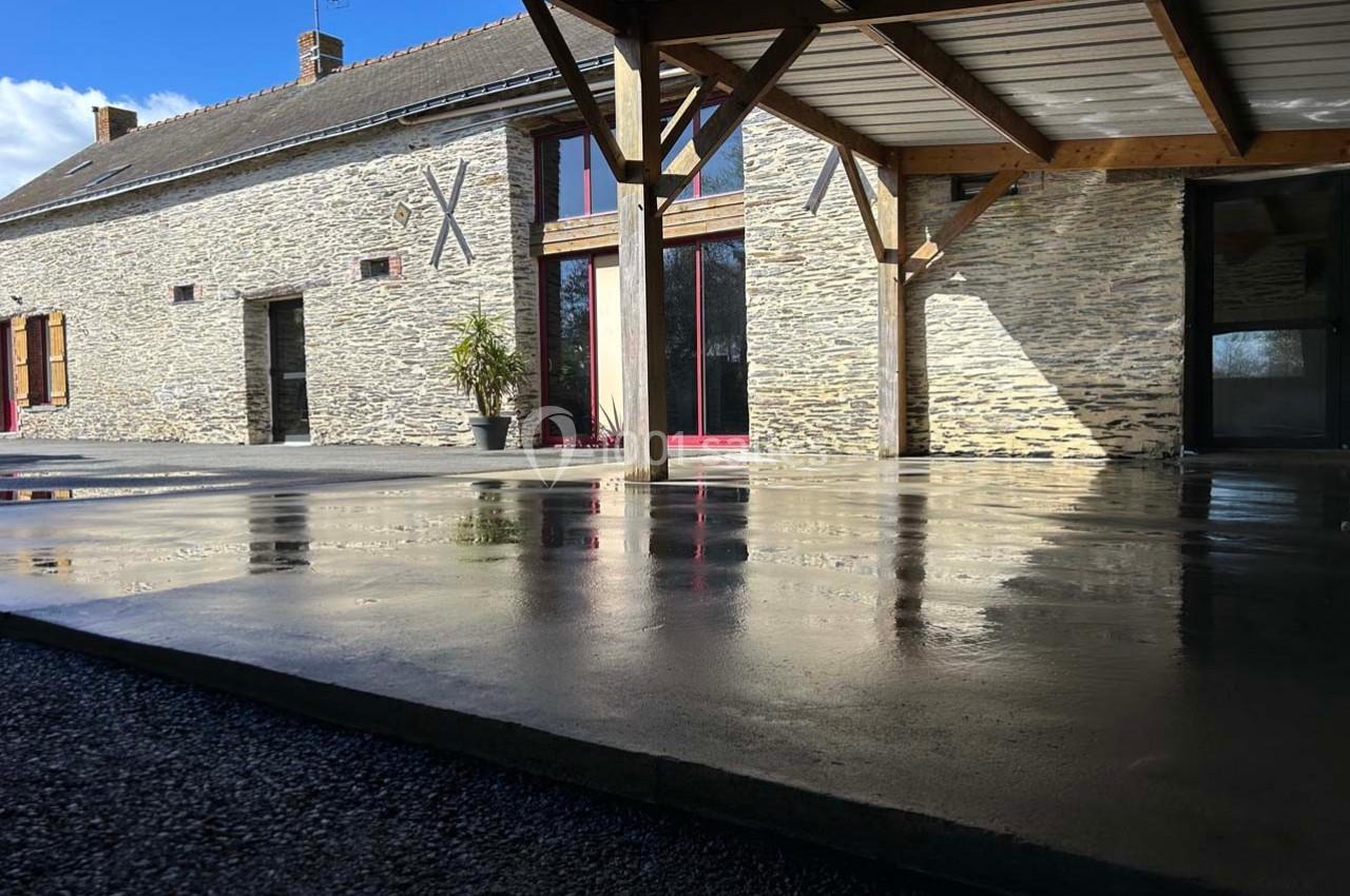 Cour en béton lisse devant une maison en pierre avec charpente en bois et grandes baies vitrées.