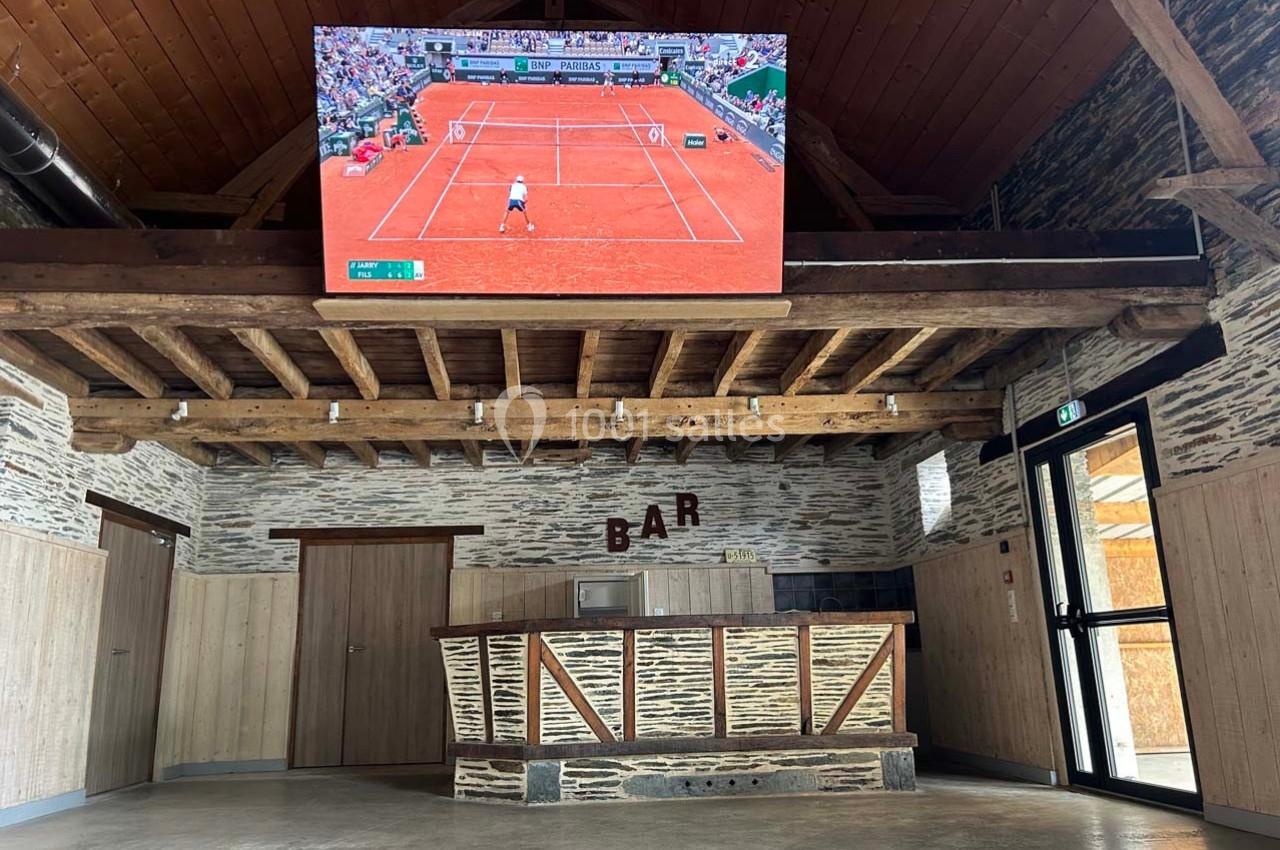 Un bar en bois avec un écran diffusant un match de tennis sur terre battue, dans un espace à plafond en poutres apparentes.