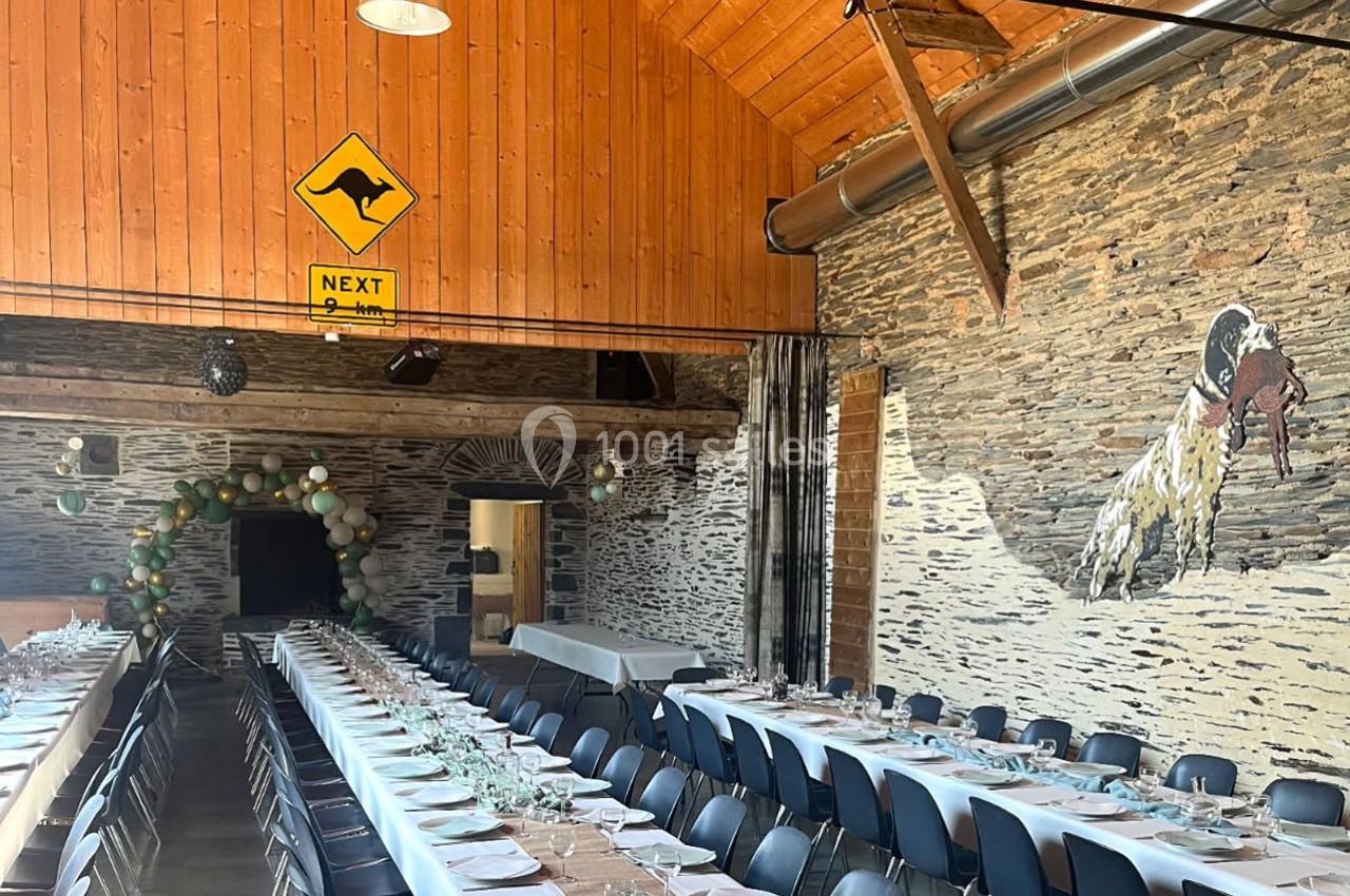 Salle de réception avec longues tables dressées, murs en pierre, décoration de ballons et panneau jaune avec un kangourou.